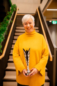 Louise is in a yellow jumper holding the tabbron award with both hands. she has short white hair and is smiling at the camera. 